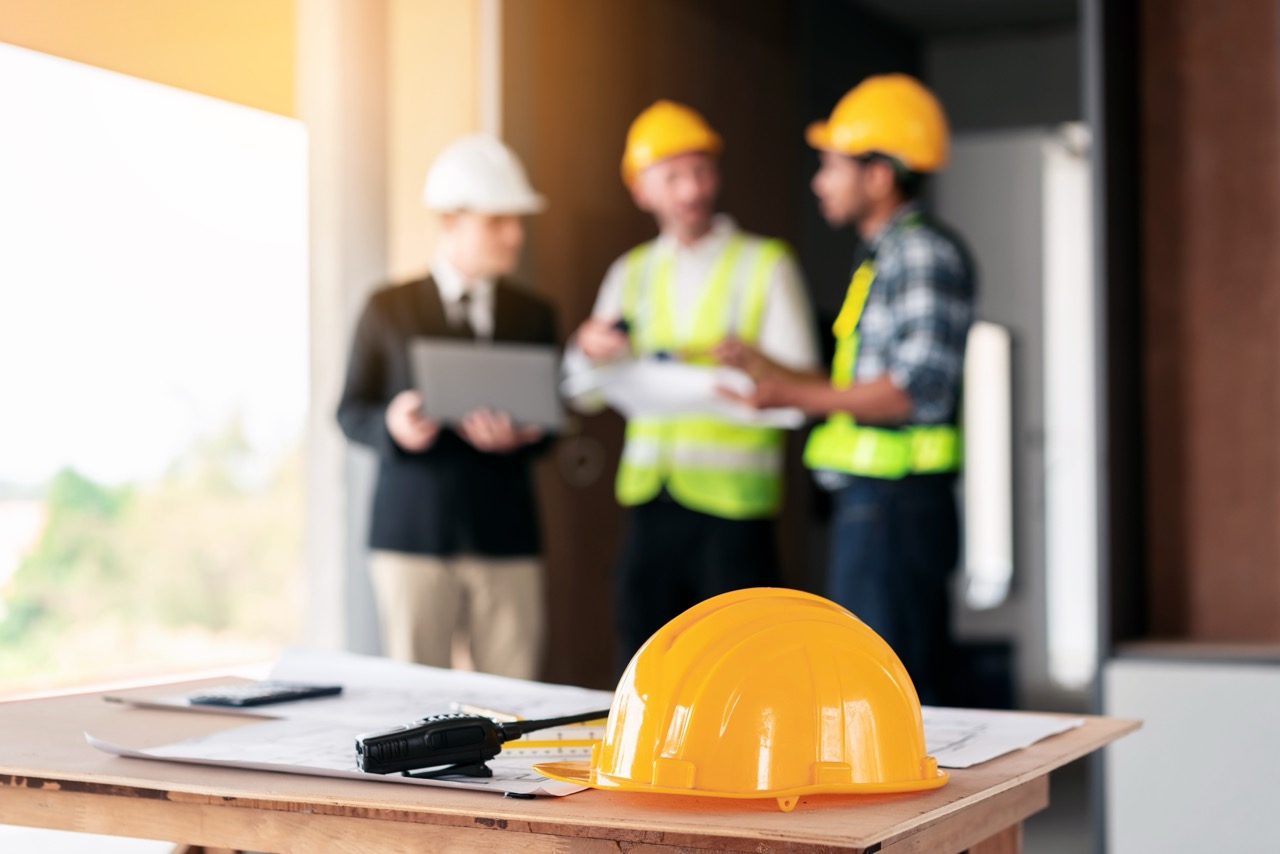 Three men wearing hard hats are standing around a table with blueprints and a yellow hard hat